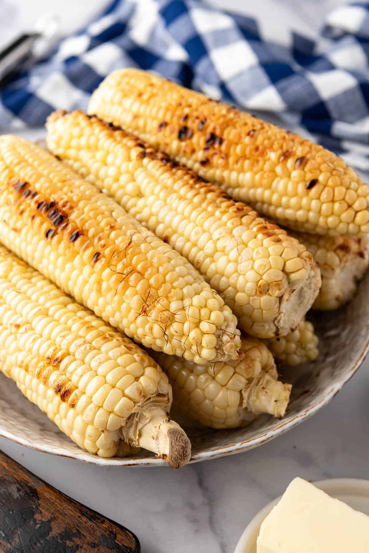 An image of grilled corn on the cob on a serving platter with a blue checkered cloth in the background.