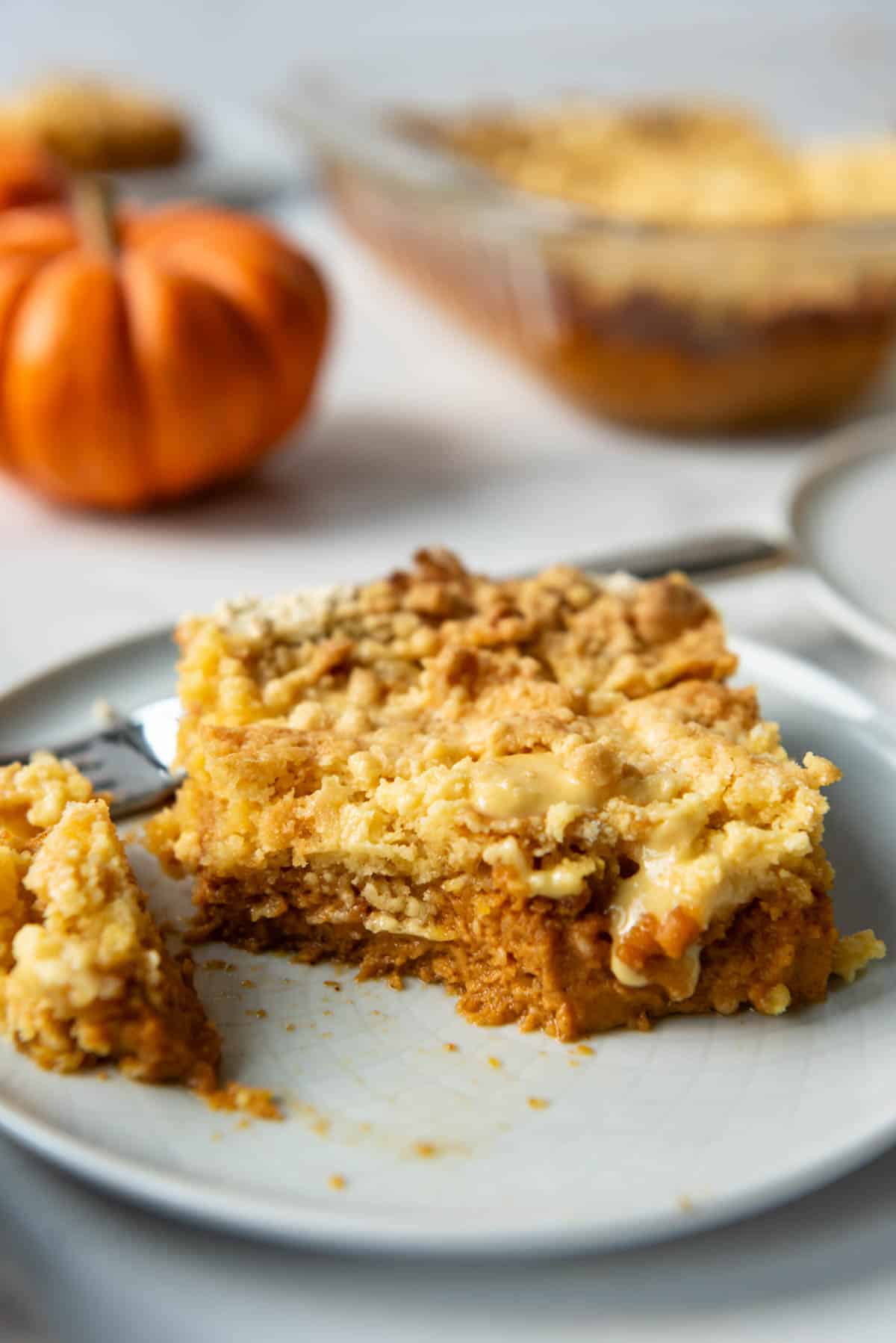 A slice of pumpkin dump cake with a bite taken out of it on a white plate with a mini orange pumpkin in the background.