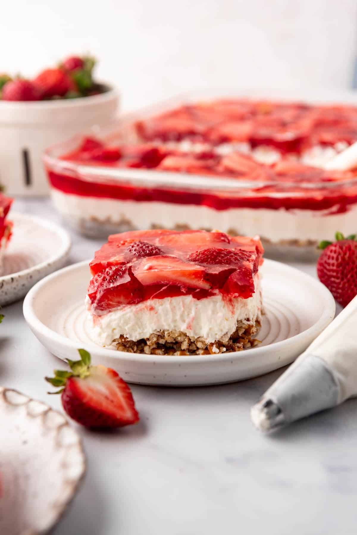 A slice of strawberry pretzel salad dessert on a plate in front of the pan of jello salad.