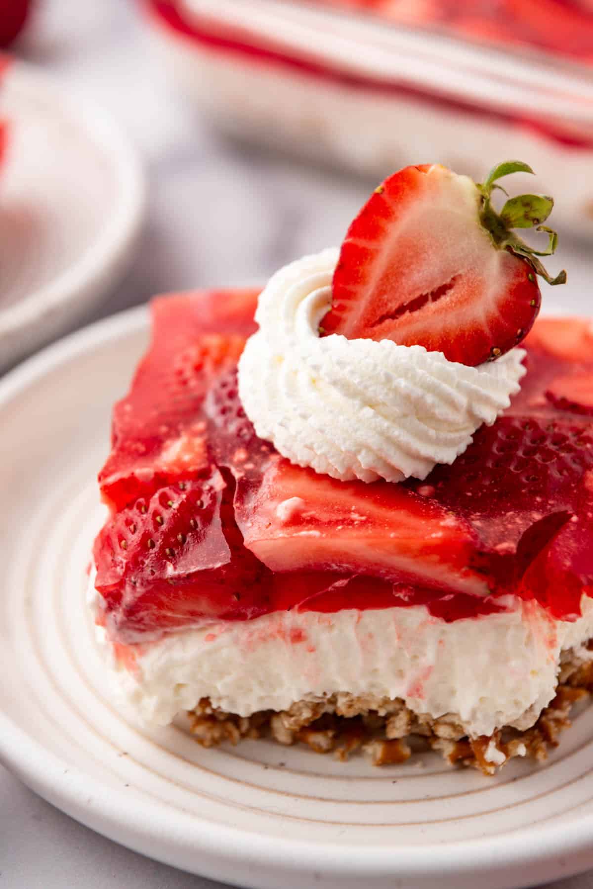 A serving of strawberry pretzel salad decorated with a swirl of whipped cream and half a strawberry on a plate.