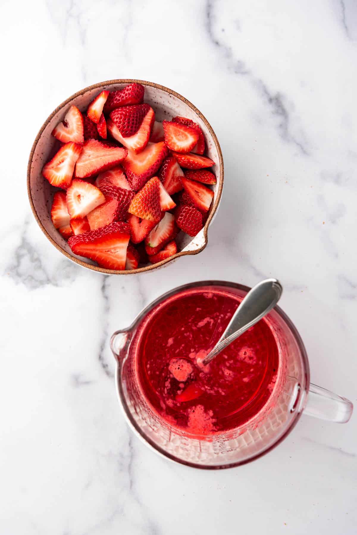 Sliced strawberries in a bowl next to dissolved strawberry jello in a glass mixing bowl with a spoon.