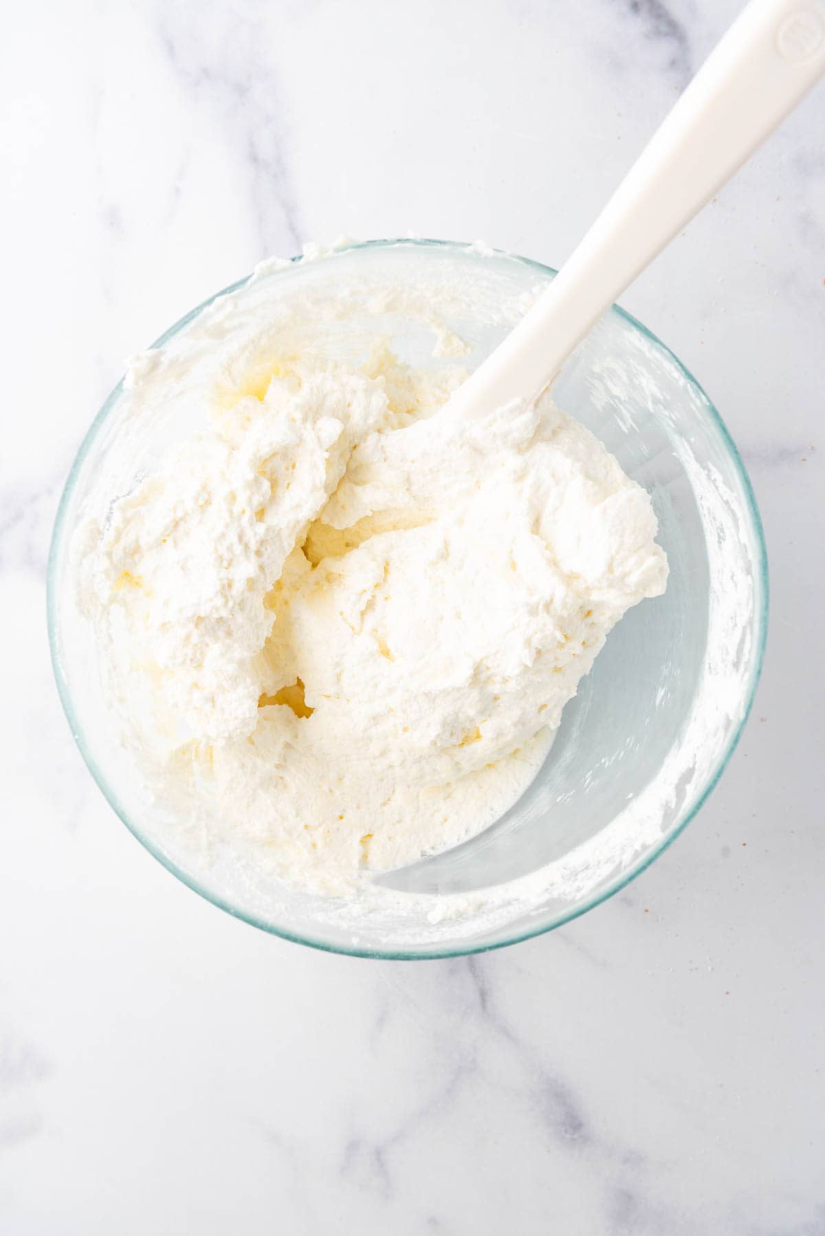 Combining cream cheese and whipped cream in a glass mixing bowl with a white spatula.