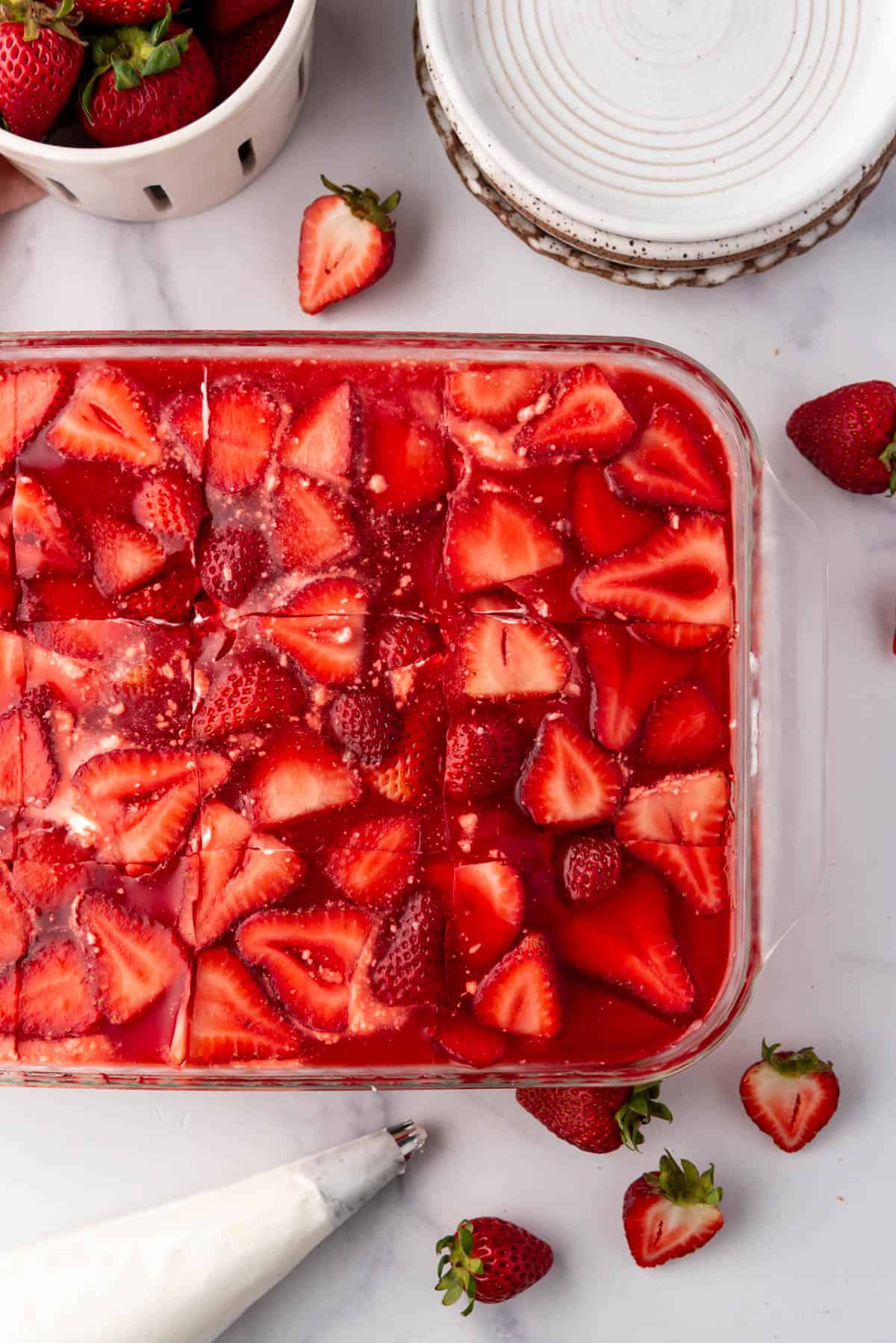 An overhead image of a strawberry pretzel jello salad next to whipped cream in a piping bag and strawberries.