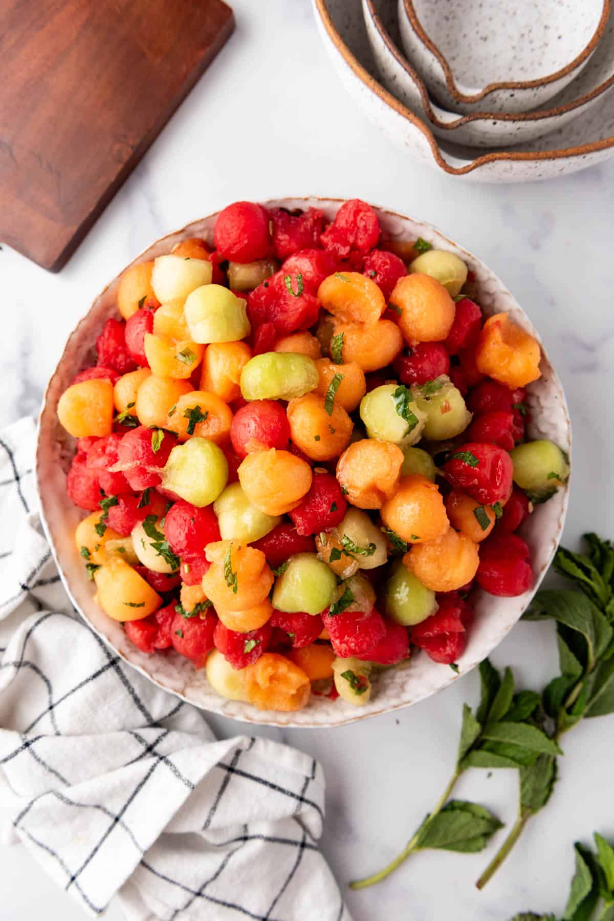 An overhead image of a bowl of melon salad with melon balls surrounded by a wooden cutting board, bowls, and a cloth napkin.