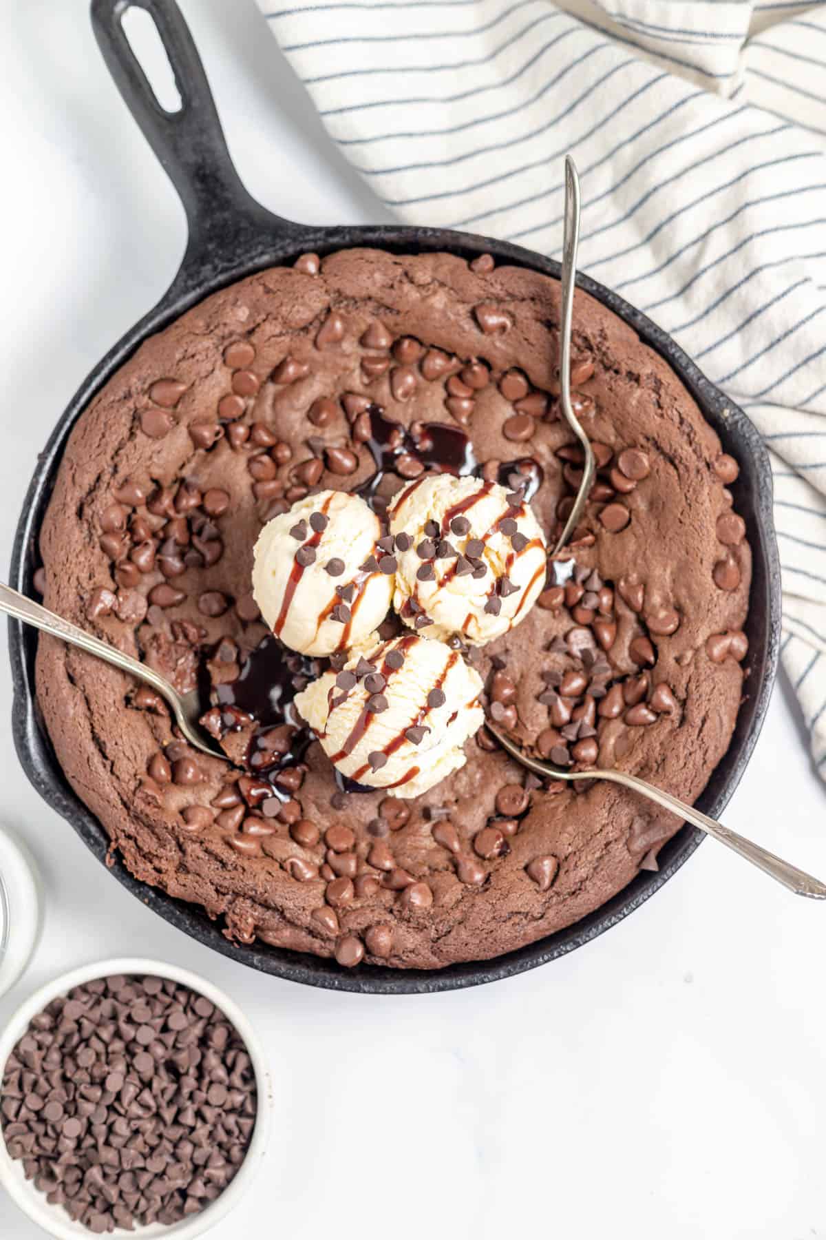 An overhead image of a chocolate skillet cookie with three spoons in it and ice cream on top.