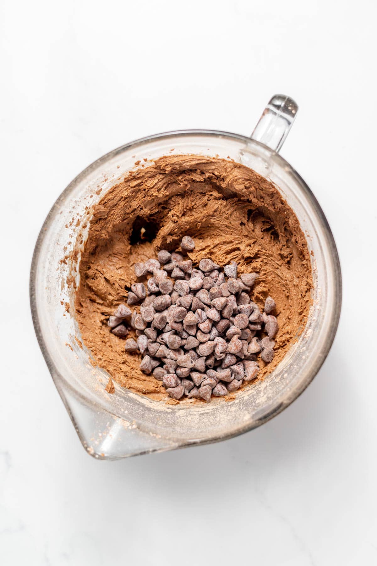 Adding chocolate chips to chocolate cookie dough in a large glass mixing bowl.