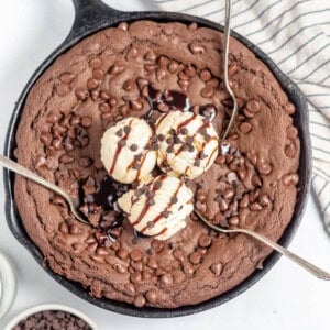 An overhead image of a chocolate skillet cookie with three spoons in it and ice cream on top.