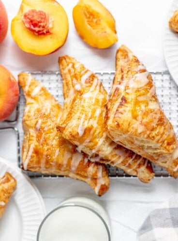 Three peach turnovers on a wire rack next to a glass of milk and fresh peaches.