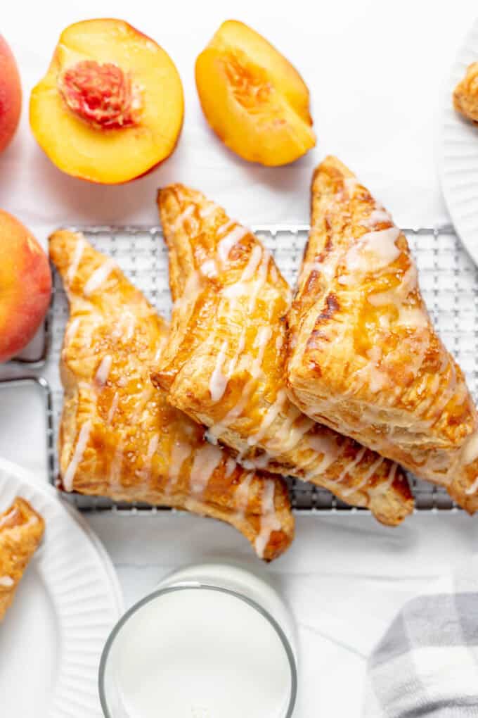 Three peach turnovers on a wire rack next to a glass of milk and fresh peaches.