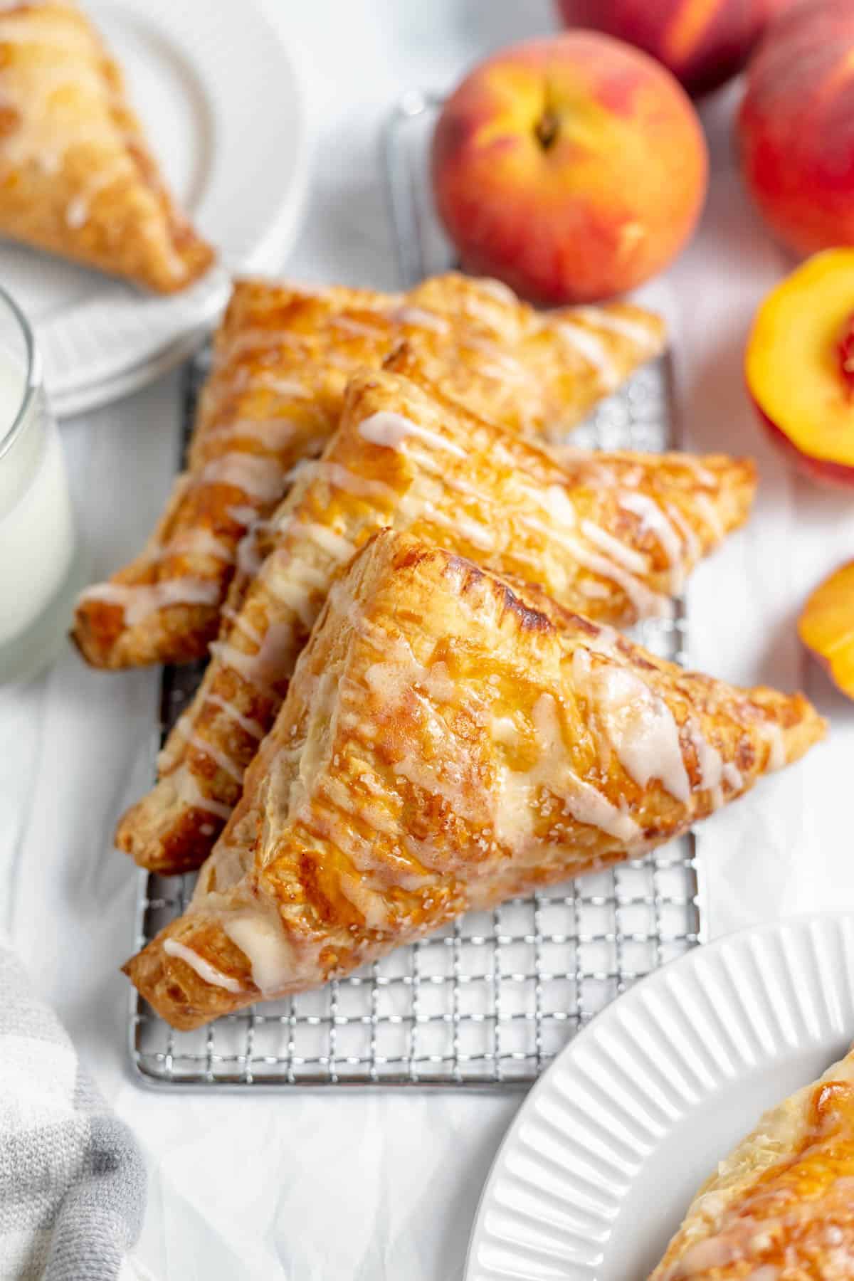 Three glazed peach turnovers on a wire rack next to fresh  peaches.