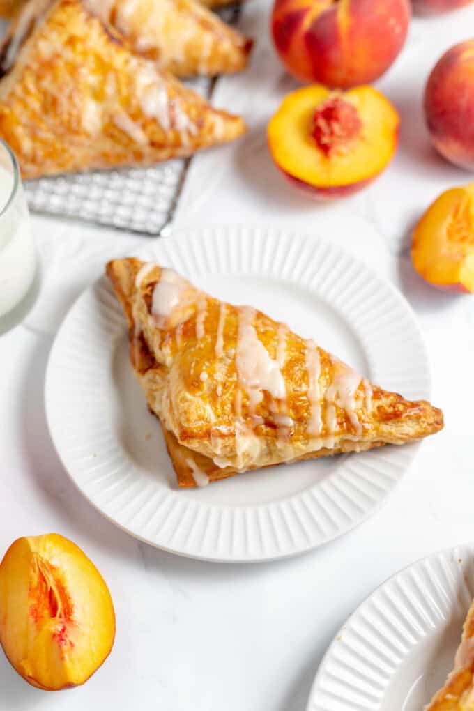 An overhead image of a peach turnover on a white plate next to fresh peaches and more turnovers.