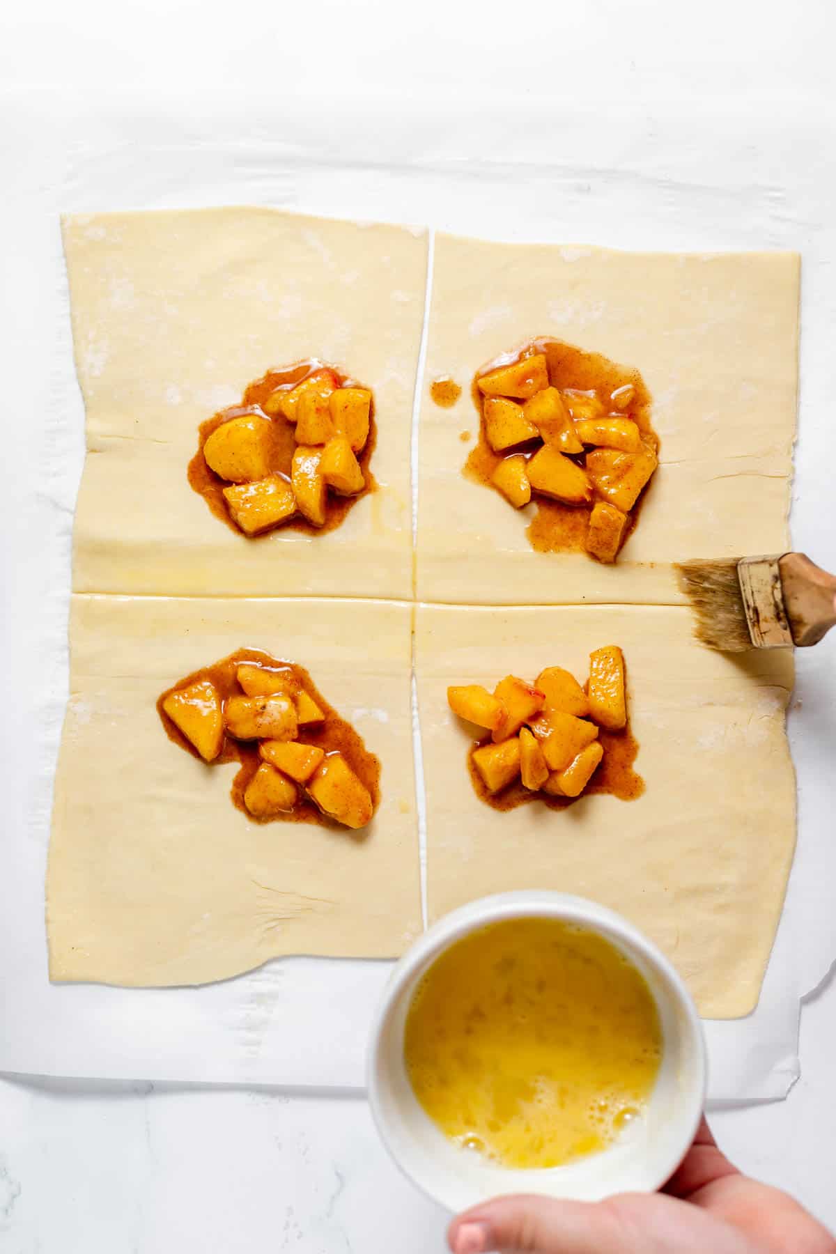 Adding peach filling and brushing edges of puff pastry dough with egg wash before sealing.