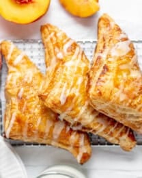 Three peach turnovers on a wire rack next to a glass of milk and fresh peaches.