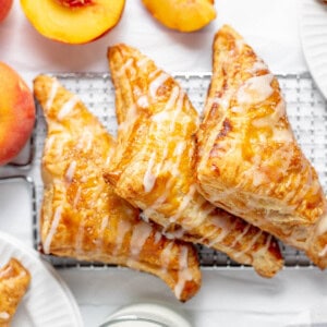 Three peach turnovers on a wire rack next to a glass of milk and fresh peaches.