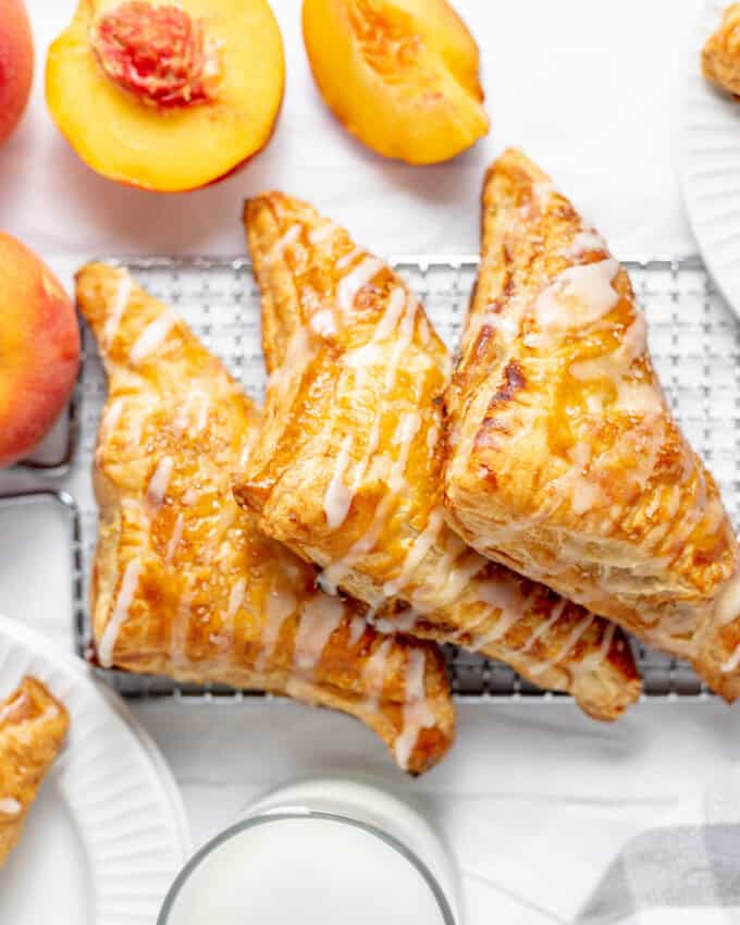 Three peach turnovers on a wire rack next to a glass of milk and fresh peaches.