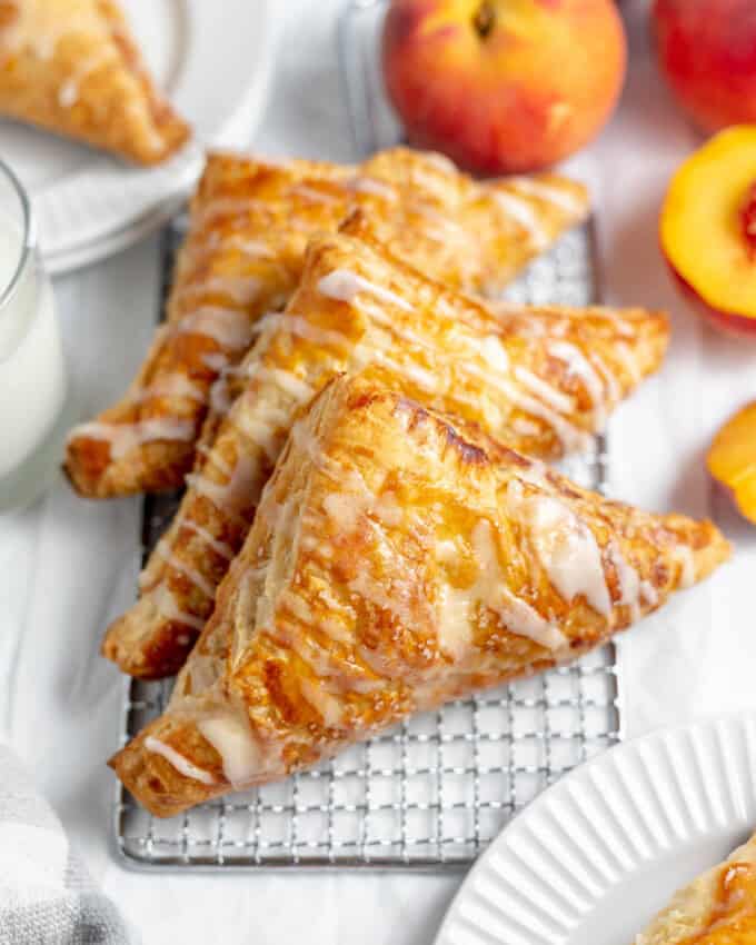 Three glazed peach turnovers on a wire rack next to fresh peaches.