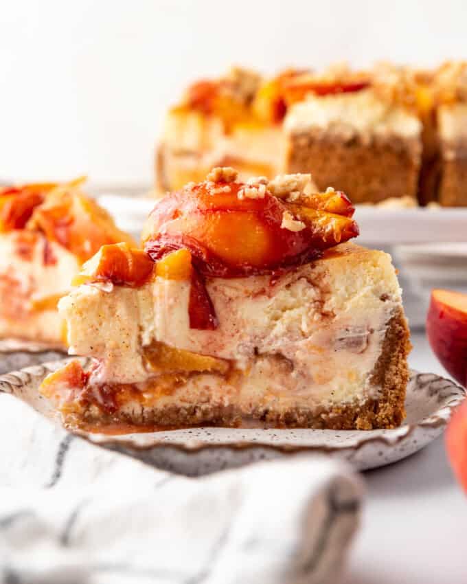 A slice of peach cobbler cheesecake on a plate with roasted fresh peaches on top.