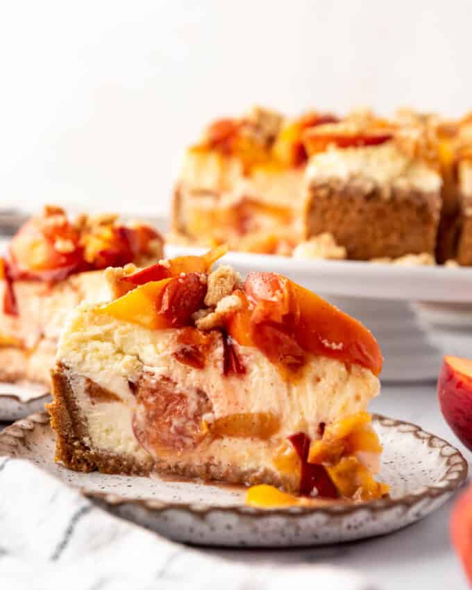 A side angle of a slice of peach cobbler cheesecake on a plate in front of the rest of the cheesecake on a cake stand.