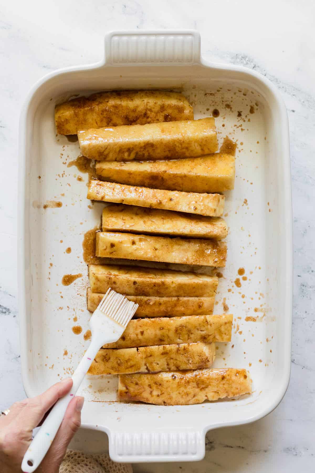 Brushing pineapple spears with melted butter and sprinkling them with brown sugar and cinnamon in a white baking dish.