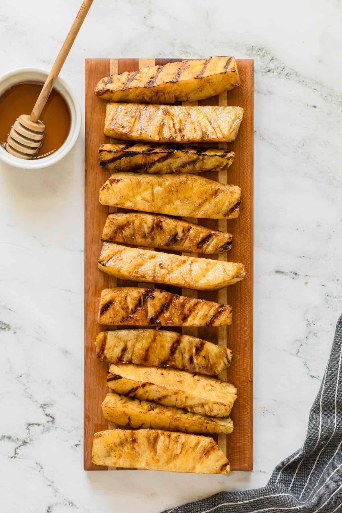 An overhead image of grilled pineapple spears on a wooden cutting board next to a small bowl of honey.