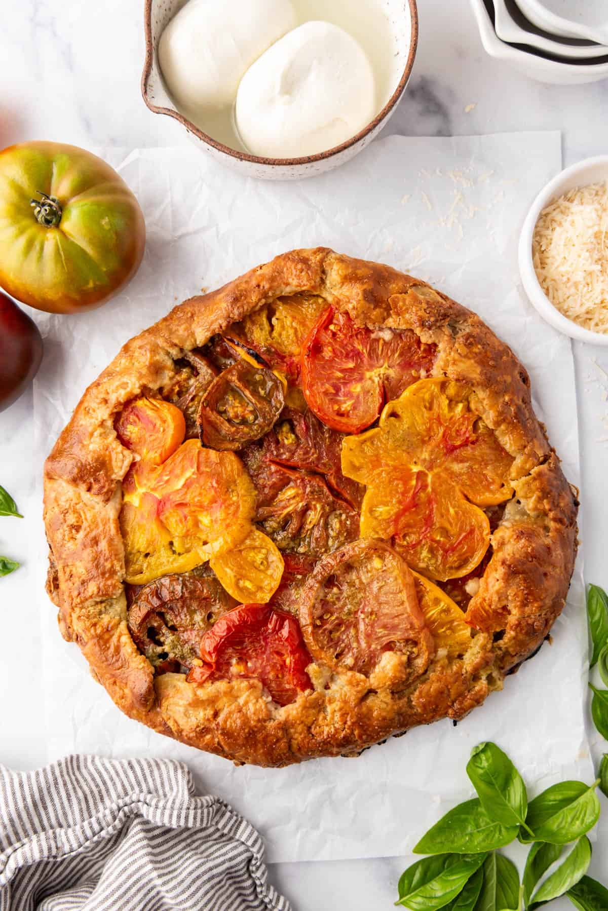 Une image d'une galette de tomates cuite à côté d'un linge rayé, de tomates héritage et de boules de burrata.