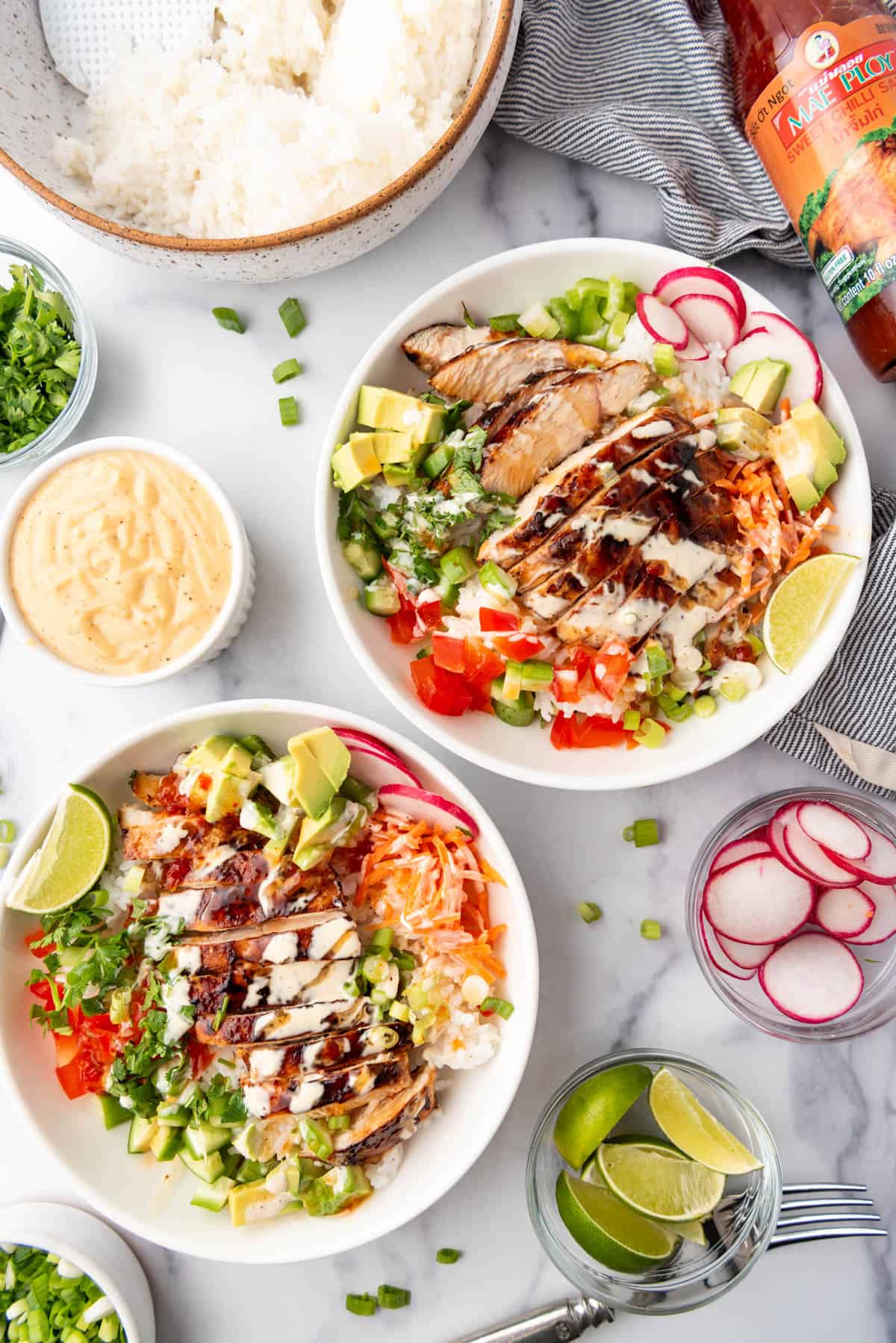 Two white bowls filled with sticky rice, chopped veggies, huli huli chicken, and covered in sweet chili sauce and yum yum sauce surrounded by small bowls of toppings.