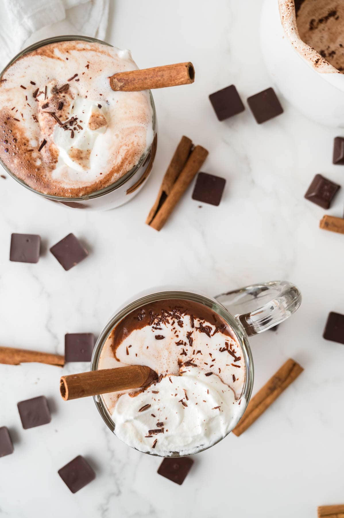 An overhead image of two mugs of hot chocolate surrounded by cinnamon and squares of dark chocolate.