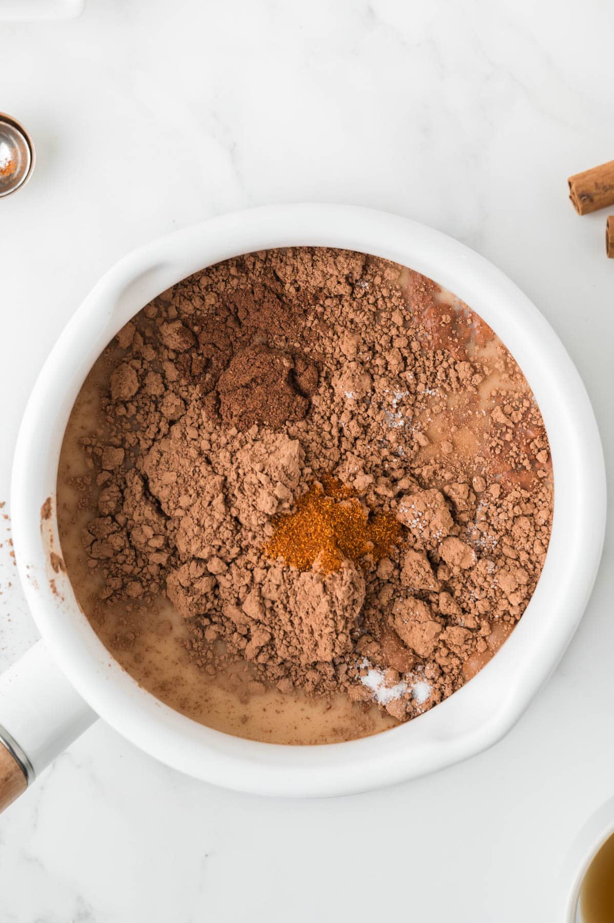 Combining milk, cocoa powder, cinnamon, and cayenne pepper in a white pot.