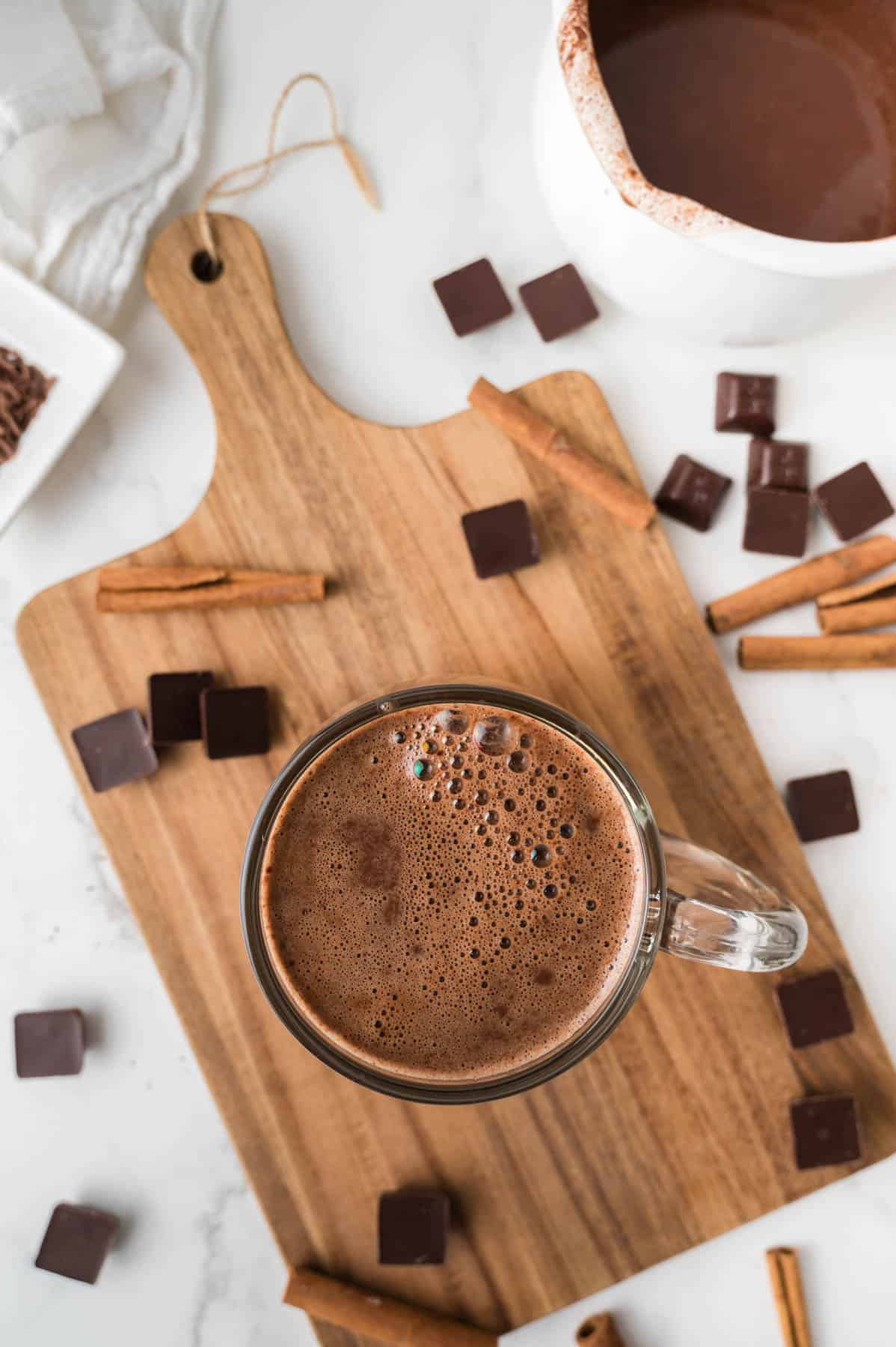 An overhead image of a mug of Mexican hot chocolate surrounded by dark chocolate squares and cinnamon sticks on a wooden cutting board.