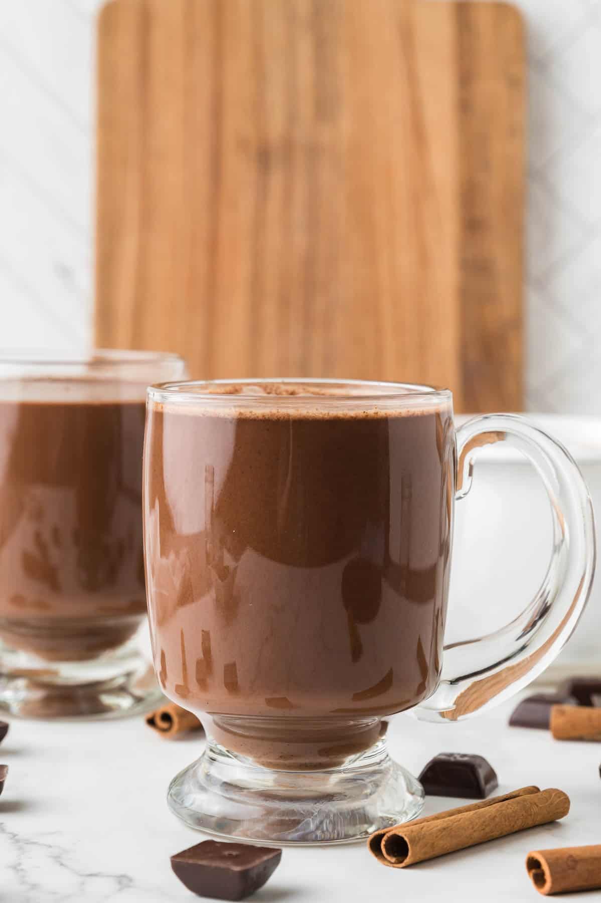 Two glass mugs of Mexican hot chocolate with cinnamon sticks nearby.