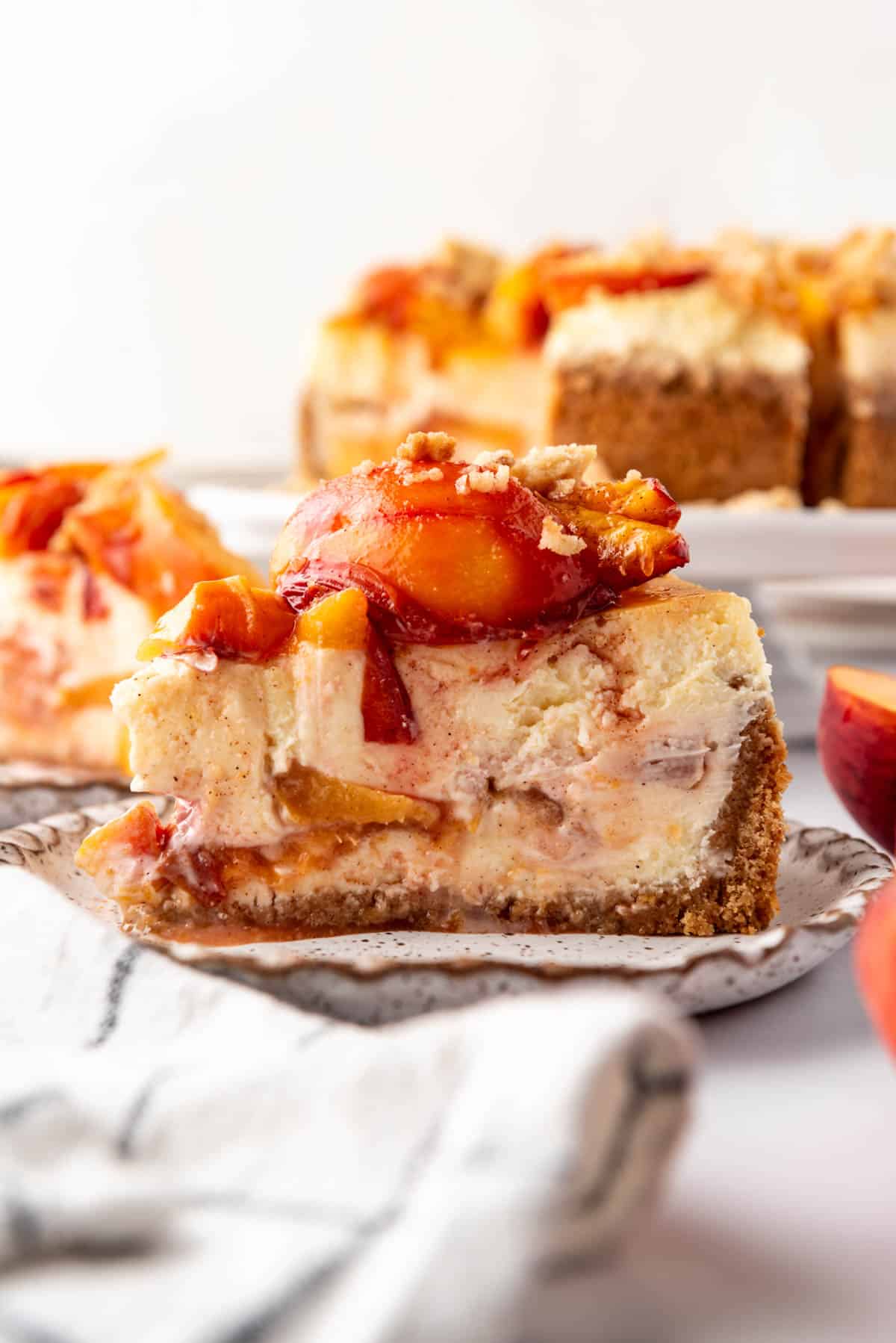 A slice of peach cobbler cheesecake on a plate with roasted fresh peaches on top.