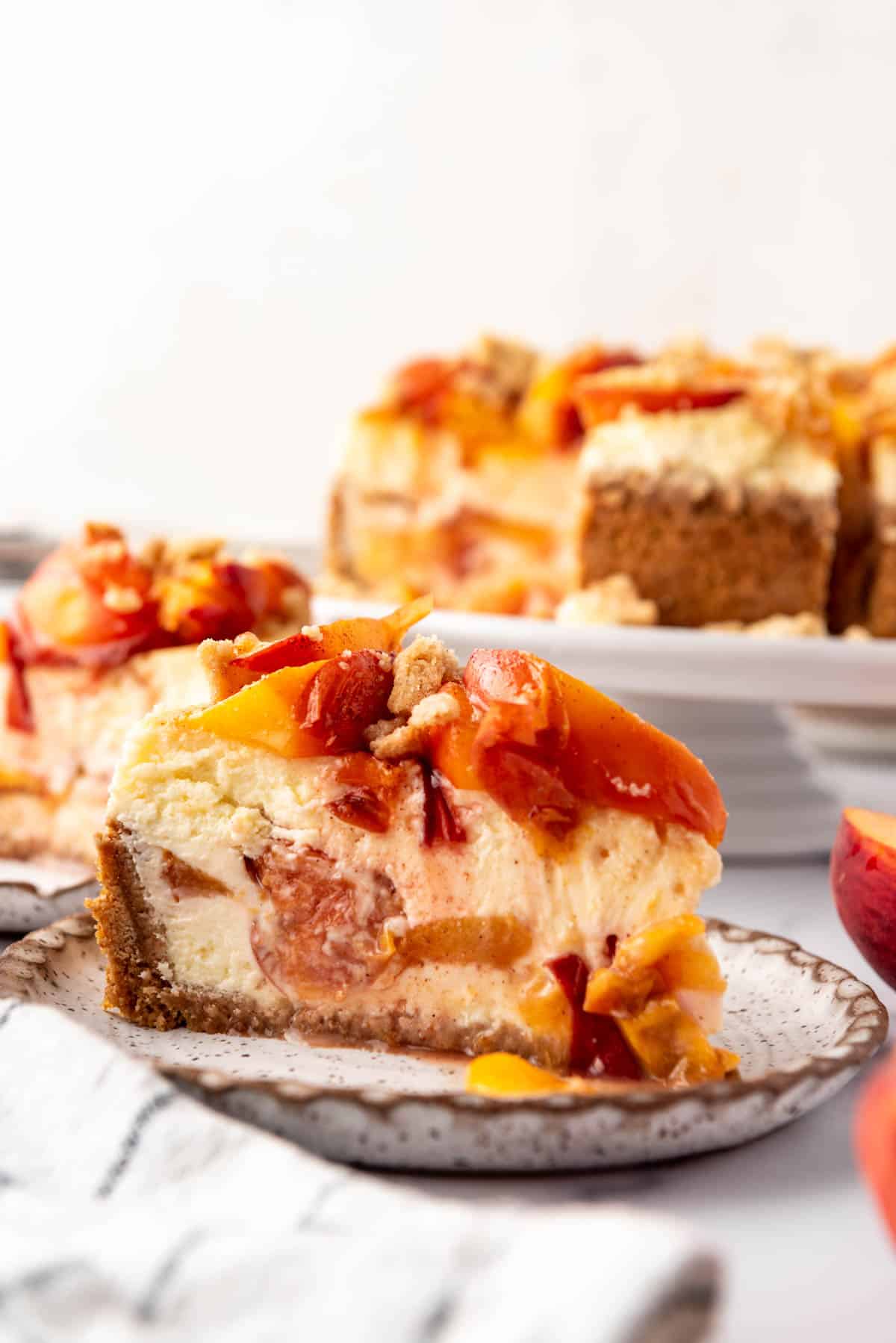 A side angle of a slice of peach cobbler cheesecake on a plate in front of the rest of the cheesecake on a cake stand.