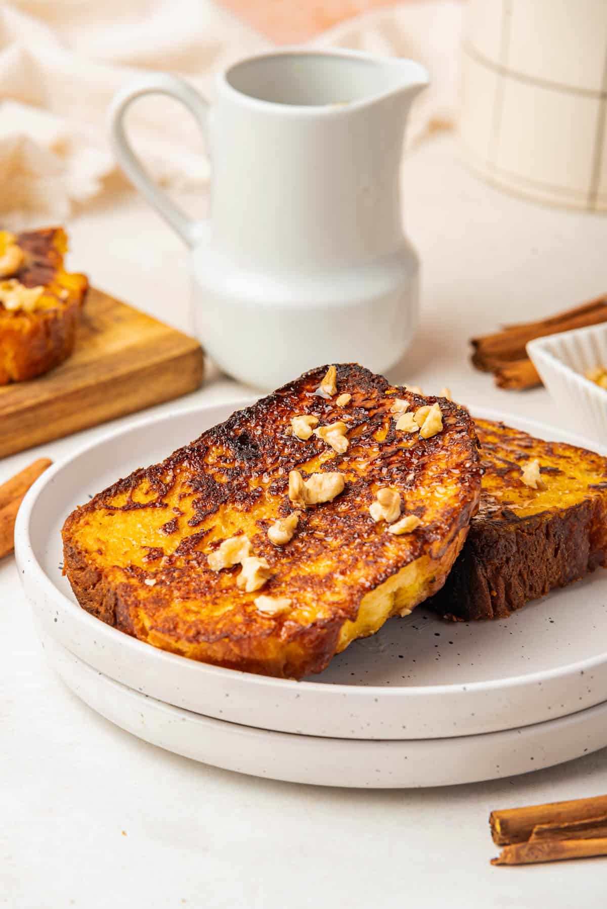 Slices of pumpkin french toast on two nested white plates with a white pitcher behind them.