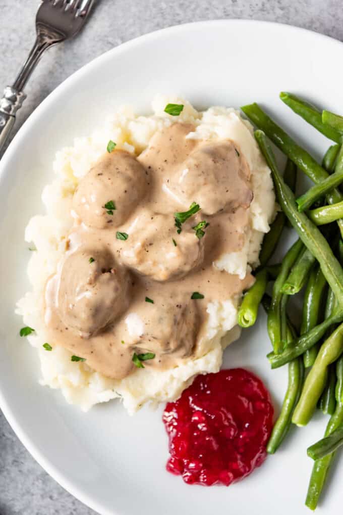 An overhead image of Swedish meatballs served over mashed potatoes with green beans and lingonberry jam on a white plate.