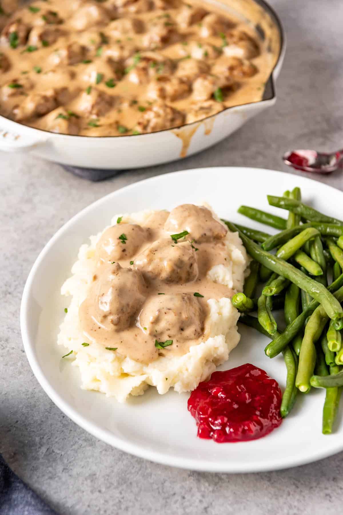 An image of a plate of Swedish meatballs served over mashed potatoes with the sauce, lingonberry jam, and green beans.