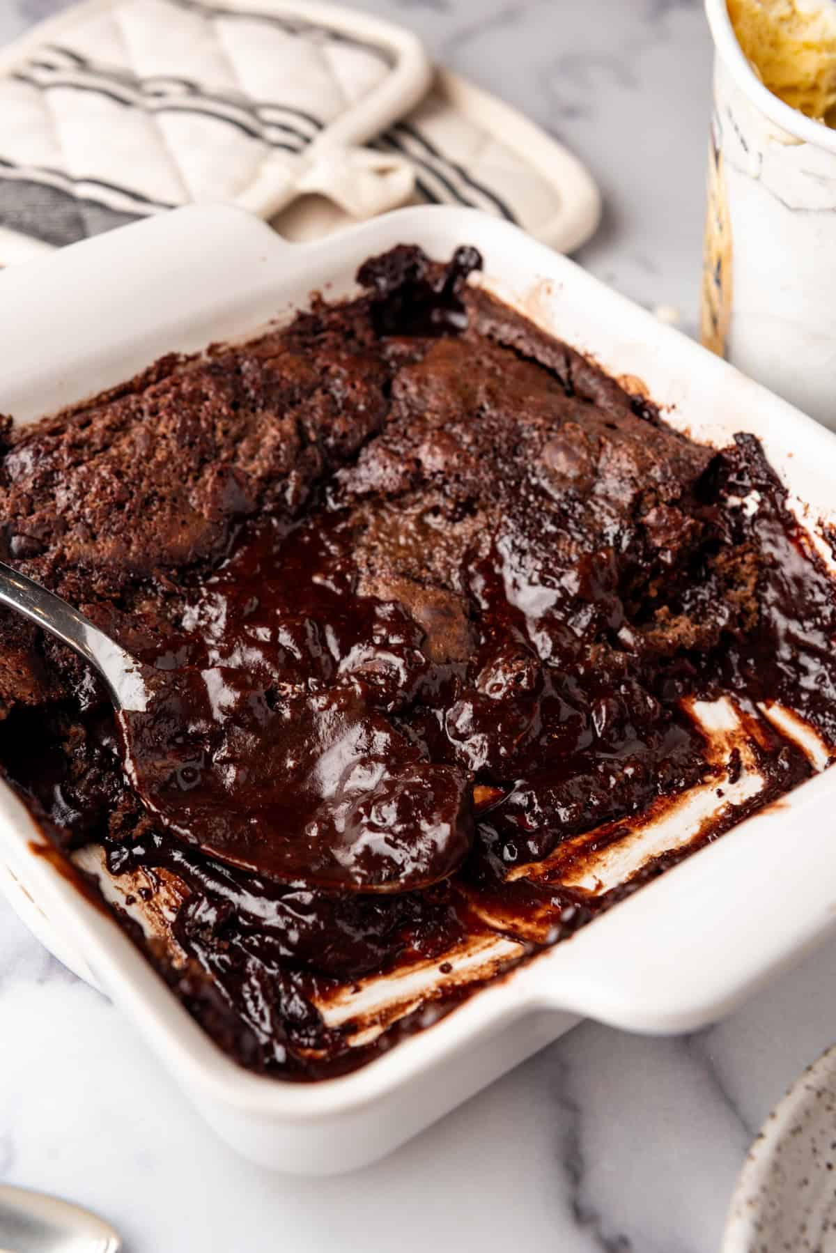 A large serving spoon in a white baking dish of chocolate cobbler cake.