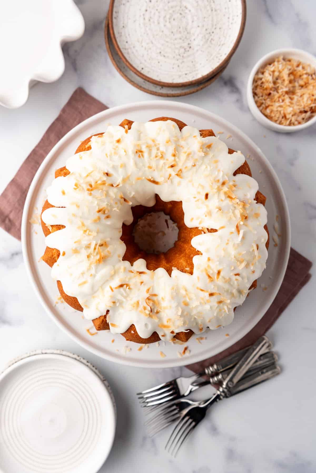 A decorated coconut bundt cake on a white cake plate surrounded by forks and plates.