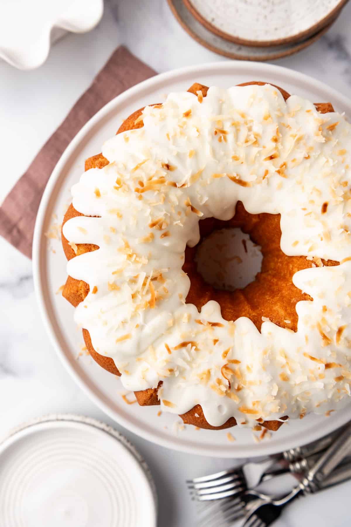 A decorated coconut bundt cake on a white cake plate surrounded by forks and plates.