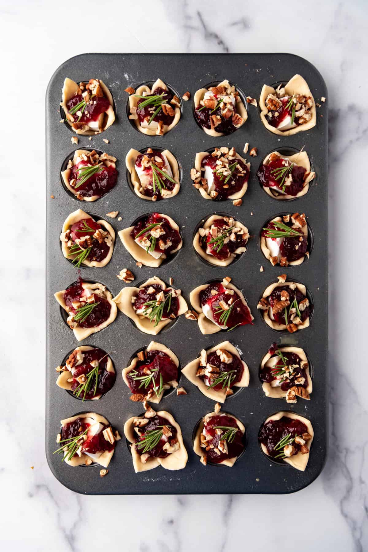 Adding cranberry sauce, chopped pecans, and rosemary sprigs to make cranberry brie bites appetizers.