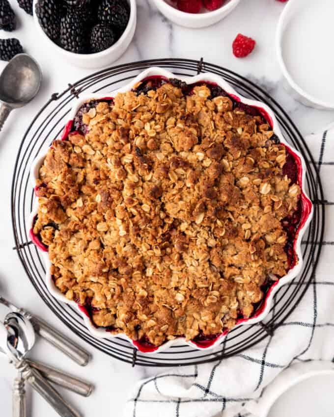 A baked mixed berry crisp cooling on a wire rack next to bowls of raspberries and blackberries.