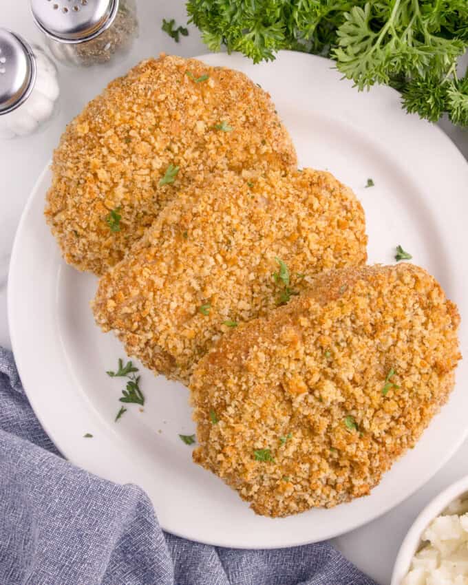 An overhead image of cooked shake n bake pork chops on a white plate next to a bowl of mashed potatoes.