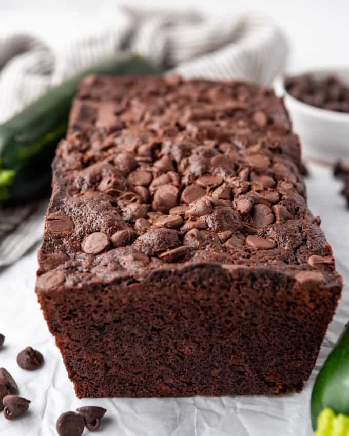 A loaf of chocolate zucchini bread on a white surface with zucchini and chocolate chips around it.