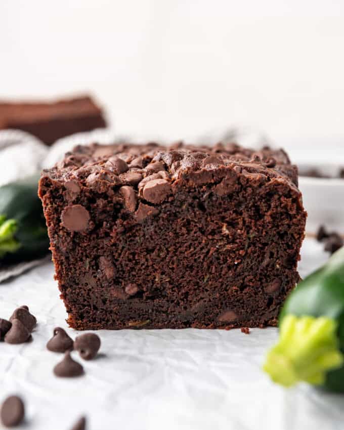 A loaf of chocolate zucchini bread on a white surface with zucchini and chocolate chips around it.