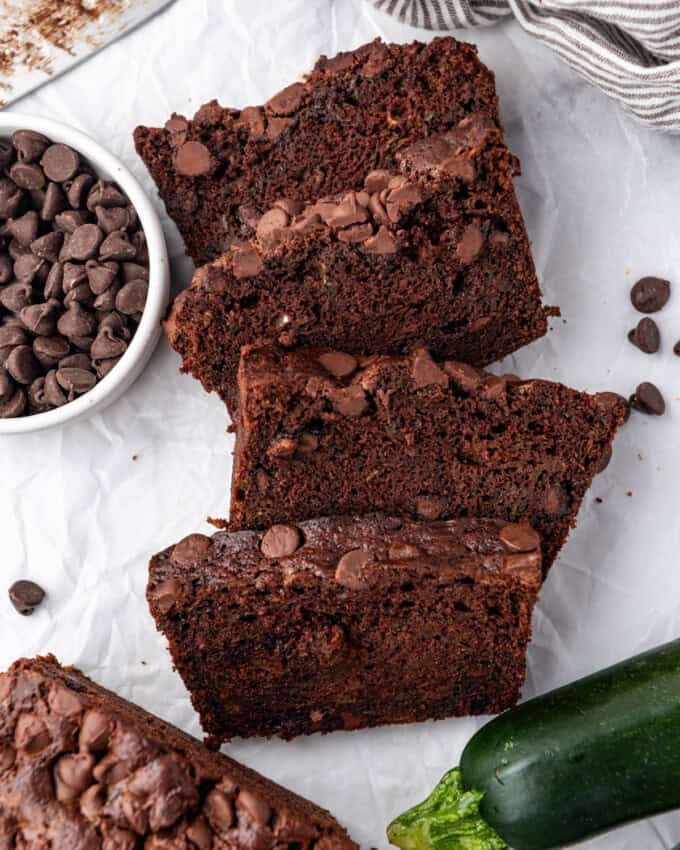 Top view of slices of chocolate zucchini bread on a white surface with zucchini and chocolate chips around them.