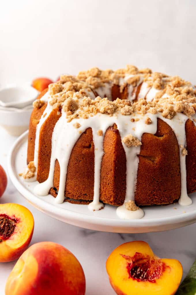 A peach pound cake with glaze and crumbles on top with fresh peaches nearby.