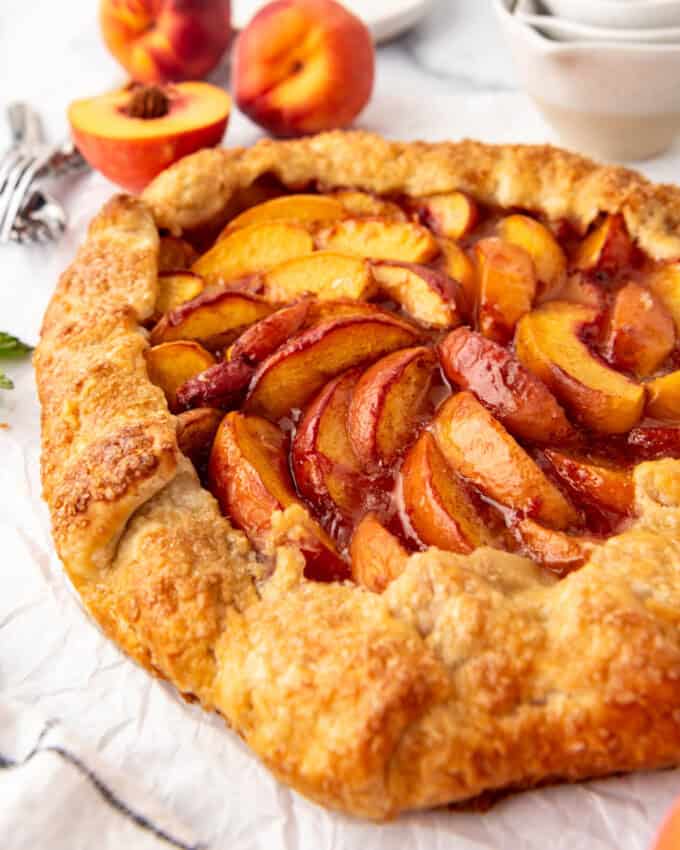 A side angle of a peach galette in front of fresh peaches and measuring cups.