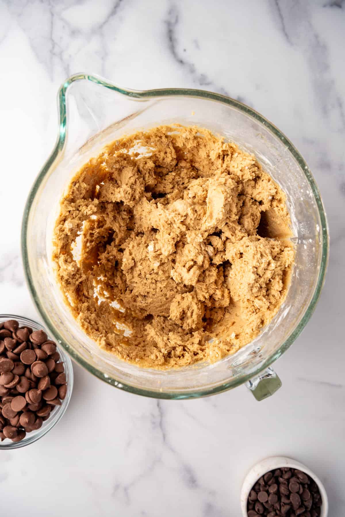 Oatmeal peanut butter cookie dough in a glass mixing bowl surrounded by chocolate chips.