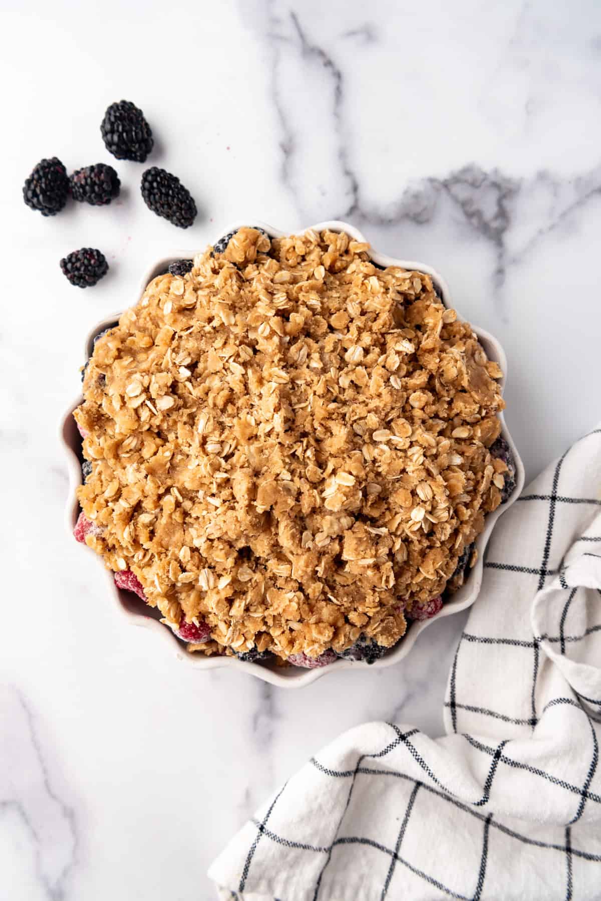 Adding an oat crisp topping to a berry dessert on a white surface  next to blackberries and a checked cloth.