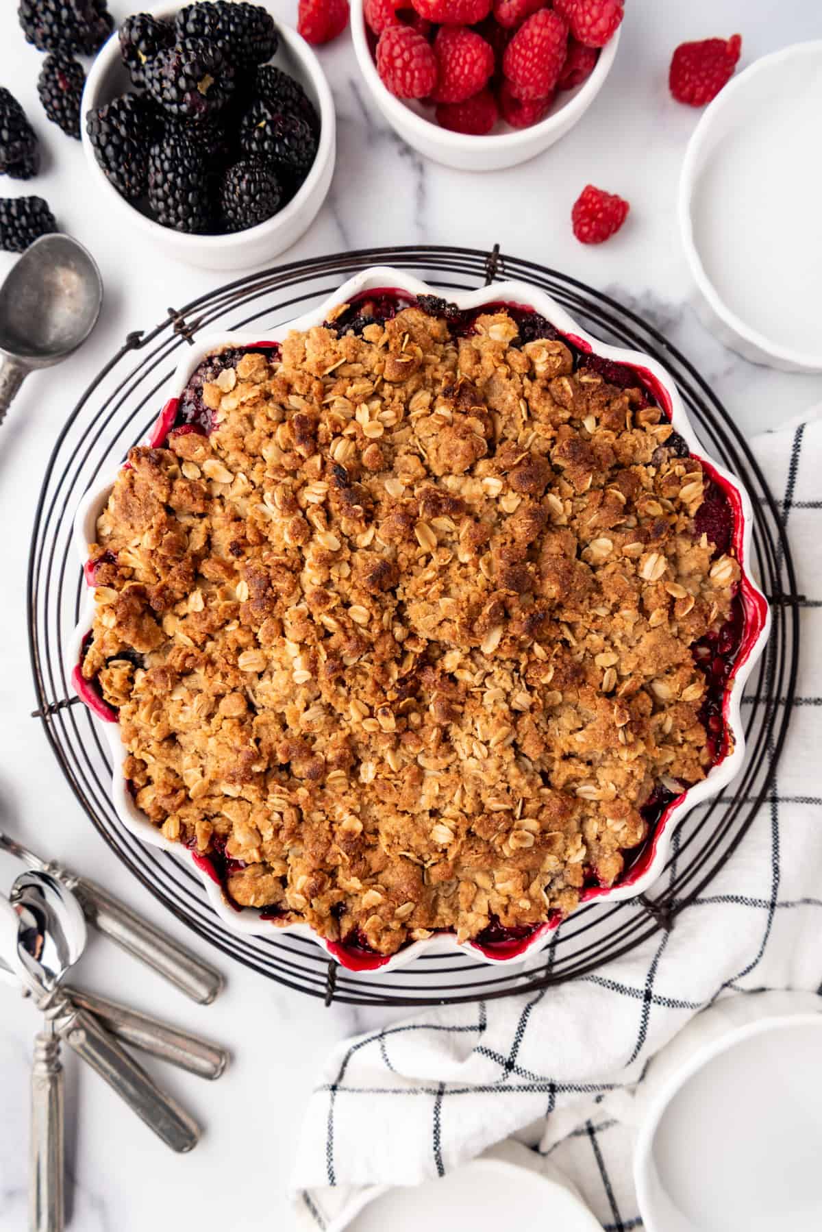 A baked mixed berry crisp cooling on a wire rack next to bowls of raspberries and blackberries.