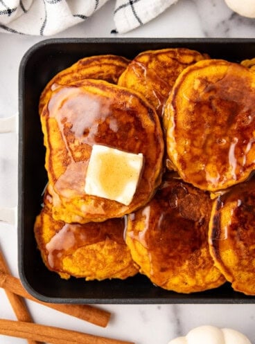 An overhead image of pumpkin pancakes with cinnamon syrup and butter on top next to cinnamon sticks and butter.